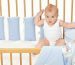 Cute baby sits curiously in a wooden baby bassinet with blue bumpers.